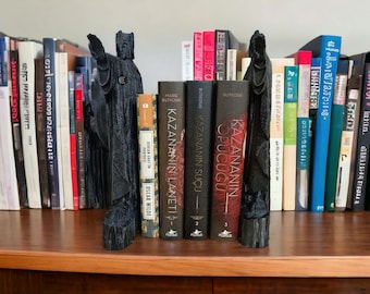 May include: Two black bookends shaped like figures in long robes stand on a bookshelf between rows of books. The bookends are made of a dark, textured material. The books on the shelf have titles in different languages, including English and Turkish.