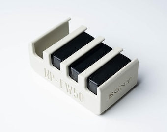 Peut inclure: Un étui de rangement pour batteries en plastique blanc avec trois batteries Sony NP-FV50 noires à l'intérieur.