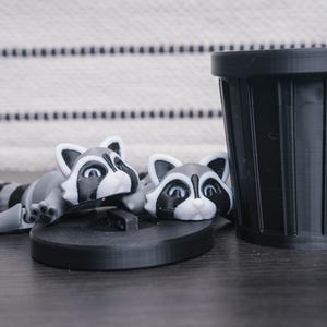 以下が含まれることがあります： 黒と白の目の黒いアライグマの置物2つと黒いゴミ箱。アライグマは横向きに寝ていて、頭はカメラの方を向いています。ゴミ箱は立っています。