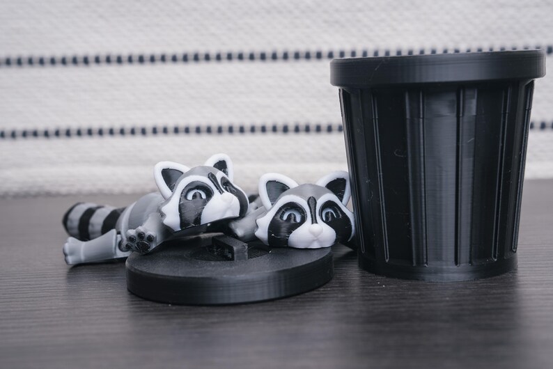 以下が含まれることがあります： 黒と白の目の黒いアライグマの置物2つと黒いゴミ箱。アライグマは横向きに寝ていて、頭はカメラの方を向いています。ゴミ箱は立っています。