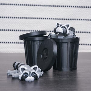 以下が含まれることがあります： ふた付きの黒いゴミ箱が2つ。1つは中にアライグマの置物が入っていて、もう1つはゴミ箱の前にアライグマの置物が床に置かれている。