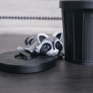 以下が含まれることがあります： 黒いプラスチック製の小さなゴミ箱の上に、アライグマの置物が置かれています。アライグマは灰色と黒で、目の周りに白い模様があります。