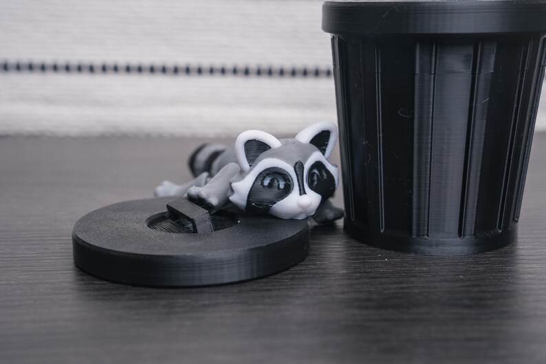 以下が含まれることがあります： 黒いプラスチック製の小さなゴミ箱の上に、アライグマの置物が置かれています。アライグマは灰色と黒で、目の周りに白い模様があります。