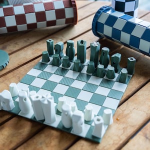 Op de afbeelding: Een groen en wit schaakbord met witte en groene schaakstukken. De schaakstukken bevinden zich in een cilindrische container met een blauw-wit geruit patroon. Er is een tweede cilindrische container met een rood-wit geruit patroon.