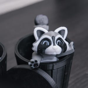 以下が含まれることがあります： 黒いゴミ箱から、グレーと白の小さなアライグマの置物が顔を出しています。アライグマは白い顔に黒い目と黒い鼻をしています。ゴミ箱はプラスチック製で、蓋が付いています。