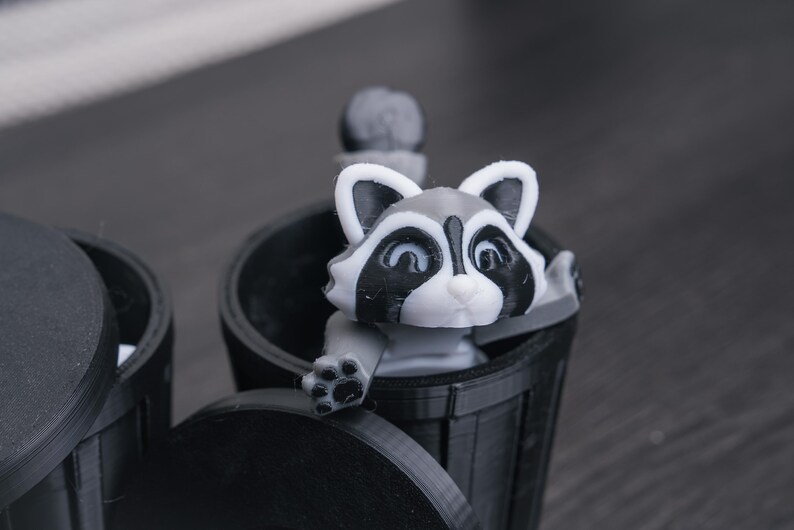 以下が含まれることがあります： 黒いゴミ箱から、グレーと白の小さなアライグマの置物が顔を出しています。アライグマは白い顔に黒い目と黒い鼻をしています。ゴミ箱はプラスチック製で、蓋が付いています。