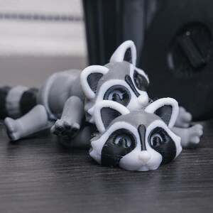 以下が含まれることがあります： グレーと白の小さなアライグマの置物2つ。左のアライグマは横向きに寝ていて、尻尾が体の周りに巻かれている。右のアライグマは腹ばいになっていて、頭が持ち上げられている。両方のアライグマは黒い目と黒い鼻をしている。