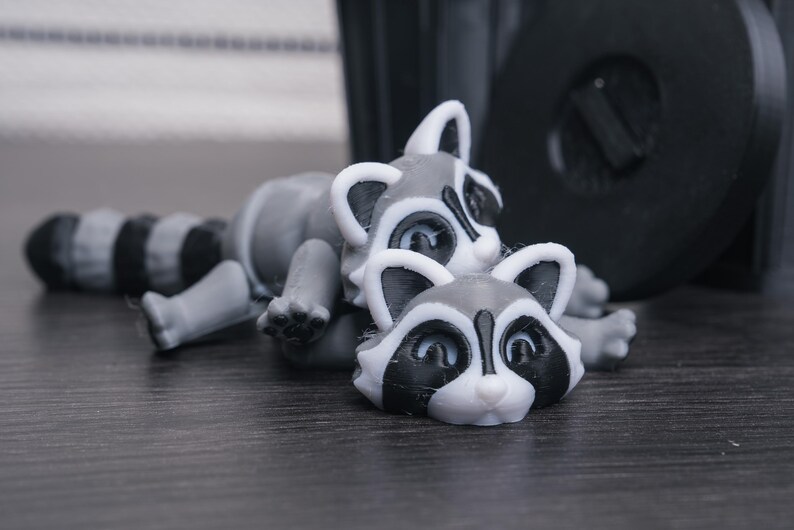 以下が含まれることがあります： グレーと白の小さなアライグマの置物2つ。左のアライグマは横向きに寝ていて、尻尾が体の周りに巻かれている。右のアライグマは腹ばいになっていて、頭が持ち上げられている。両方のアライグマは黒い目と黒い鼻をしている。