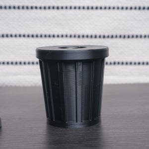 以下が含まれることがあります： リブ状のテクスチャが付いた小さな黒いプラスチック製のゴミ箱。ゴミ箱は濃い灰色の表面の上に置かれています。