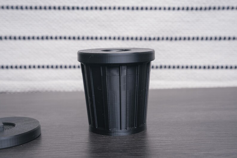 以下が含まれることがあります： リブ状のテクスチャが付いた小さな黒いプラスチック製のゴミ箱。ゴミ箱は濃い灰色の表面の上に置かれています。