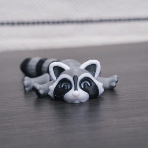 以下が含まれることがあります： 黒い目と黒い鼻を持つ、グレーと白の小さなアライグマの置物。置物は、お腹を下にして、手足を伸ばして横たわっています。