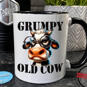 Op de afbeelding: Een witte keramische mok met een zwarte handgreep en rand, voorzien van een afbeelding van een norse koe met een ronde bril. De tekst "GRUMPY" staat boven de koe en "OLD COW" eronder. Een blauwe ovale sticker bevat de tekst "Personalise Me!!" en een zwarte stempel "HANDMADE".