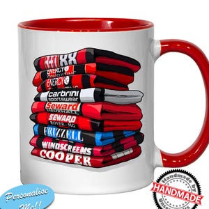 Può includere: Una tazza in ceramica bianca con bordo e manico rossi. La tazza presenta una pila di maglie da calcio rosse e nere con vari loghi di sponsor, tra cui "Energy Consulting", "Carbrini Sportswear", "Seward Vauxhall", "Seward Rover-MG", "Frizzell", "Windscreens" e "Cooper". La tazza ha anche il testo "Personalizza me!!" e un adesivo "Handmade by Unseen Gifts".