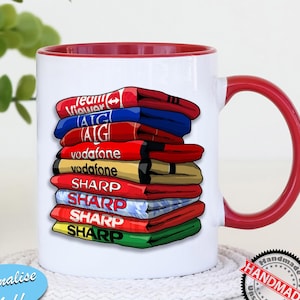 May include: A white ceramic mug with a red rim and handle. The mug features a stack of colorful soccer jerseys with the names of sponsors, including "TeamViewer", "AIG", "Vodafone", and "Sharp". The mug is on a white coaster with a "Handmade by Unseen Gifts" sticker.