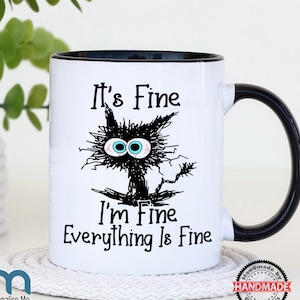 May include: White ceramic mug with a black rim and handle. The mug features a black and white illustration of a cat with the text "It's Fine I'm Fine Everything Is Fine".