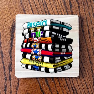 May include: A square coaster with a stack of illustrated shirts. The shirts are in various colors, including white, black, yellow, red, and blue. Each shirt has a different logo or text, such as "Kisela" and "McEwan's Lager".