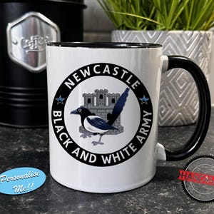 May include: White ceramic mug with a black handle and rim. The mug features a circular design with the text "NEWCASTLE BLACK AND WHITE ARMY" around a magpie illustration and a castle. A black coffee canister and a decorative plant are in the background.