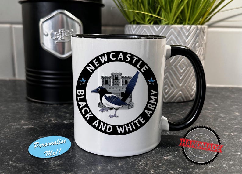 May include: White ceramic mug with a black handle and rim. The mug features a circular design with the text "NEWCASTLE BLACK AND WHITE ARMY" around a magpie illustration and a castle. A black coffee canister and a decorative plant are in the background.