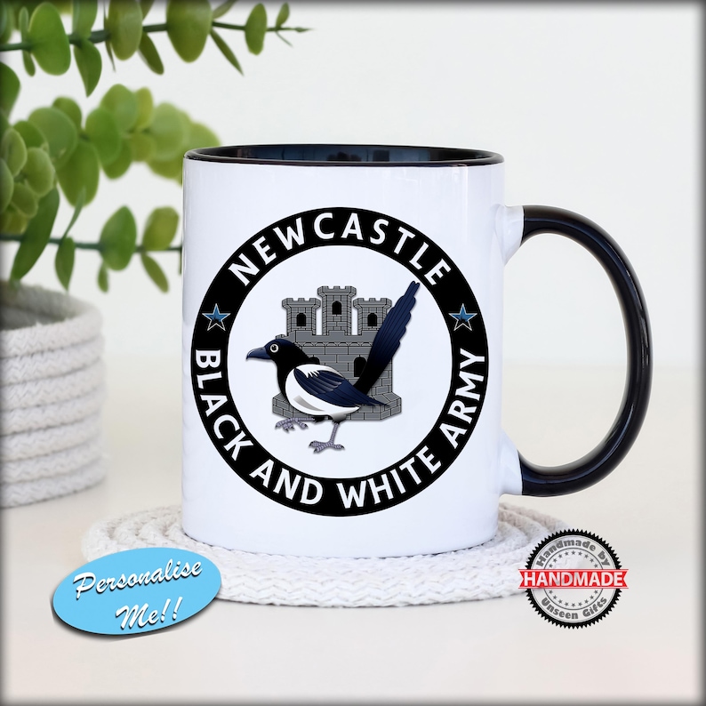 May include: White ceramic mug with a black interior and handle. The mug features a circular design with the text "NEWCASTLE BLACK AND WHITE ARMY" around a magpie and castle illustration. The mug is on a white coaster.