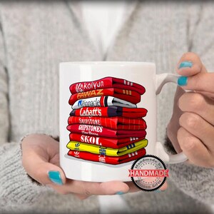 K&ouml;nnte beinhalten: Wei&szlig;e Keramiktasse mit einem bunten Design aus &uuml;bereinander gestapelten Hockeytrikots mit verschiedenen Markennamen, darunter "Kaltyn", "Fawaz", "Pinnacle", "Labatt's", "Shipstone", "Skoal" und "Handmade Gifts".