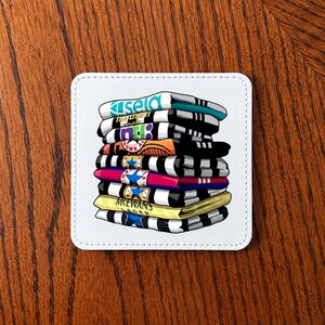 May include: A white square coaster with a stack of colorful t-shirts illustrated on it. The shirts are in various colors, including blue, black, orange, pink, and yellow, with text on each shirt. The coaster has a white border and is on a wooden surface.