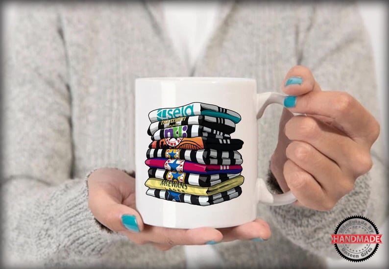 K&ouml;nnte beinhalten: Wei&szlig;e Keramiktasse mit einem bunten Stapel bedruckter Fu&szlig;balltrikots. Die Trikots sind schwarz, wei&szlig;, blau, rot, gelb und gr&uuml;n. Die Tasse hat einen schwarzen Henkel.