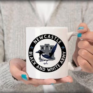 May include: White ceramic mug featuring a black and white magpie bird design with the text "Newcastle Black and White Army" in a circular emblem. The mug has a curved handle and is held by a person.
