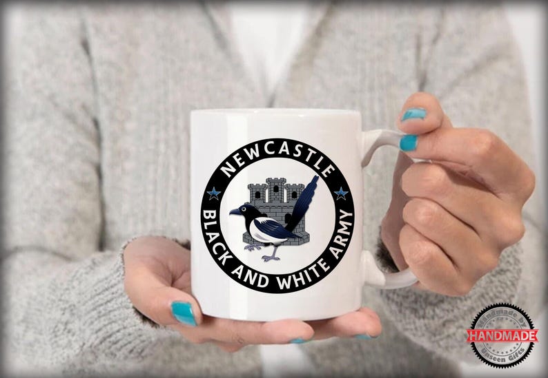 May include: White ceramic mug featuring a black and white magpie bird design with the text "Newcastle Black and White Army" in a circular emblem. The mug has a curved handle and is held by a person.