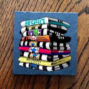May include: A stack of colorful, folded t-shirts with various logos and text, including "Kisela" and "MCEWANS LAGER". The shirts are primarily black and white striped, with accents of teal, orange, pink, and yellow. The shirts are stacked on a square, blue-gray surface.