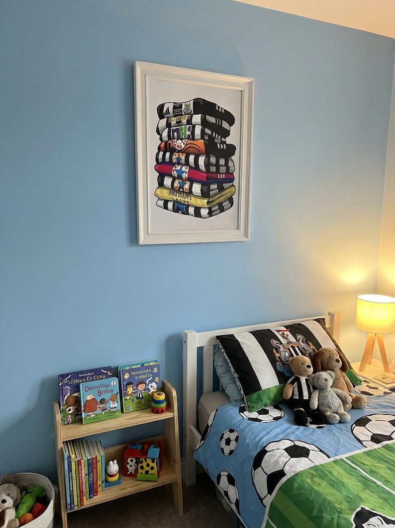 May include: A framed print of stacked jerseys hangs above a bed with a soccer-themed duvet. The print features a variety of team jerseys in black, white, yellow, and red. A small wooden bookshelf holds children's books, and stuffed animals rest on the bed.