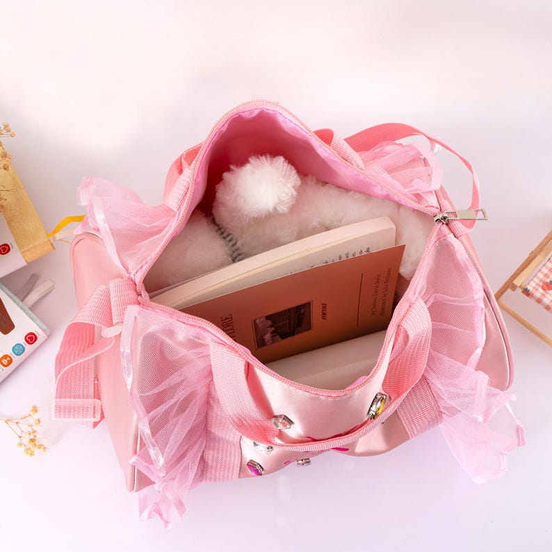 Peut inclure: Un sac &agrave; main rose avec un voile de tulle et une fermeture &eacute;clair. Le sac est ouvert, r&eacute;v&eacute;lant une peluche blanche et un livre. Le sac a des poign&eacute;es et des ornements d&eacute;coratifs. Le th&egrave;me g&eacute;n&eacute;ral est un accessoire pour enfant.