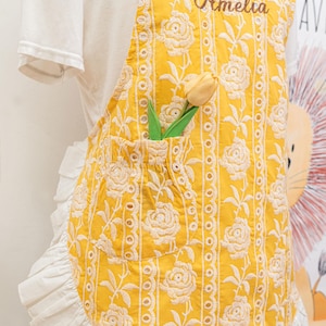 May include: Yellow apron with white floral embroidery and a pocket holding a yellow tulip. The apron has a ruffled white trim and the name "Amelia" embroidered at the top. The apron is worn over a white t-shirt.