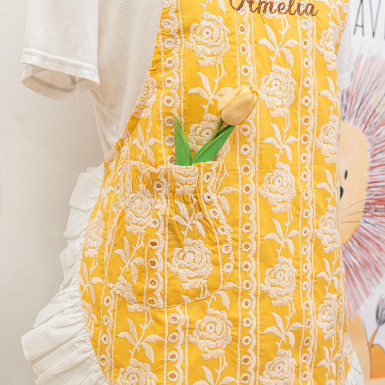 May include: Yellow apron with white floral embroidery and a pocket holding a yellow tulip. The apron has a ruffled white trim and the name "Amelia" embroidered at the top. The apron is worn over a white t-shirt.