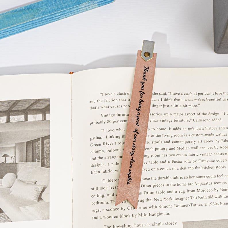 Puede incluir: Un marcap&aacute;ginas de cuero marr&oacute;n claro con el texto "Thank you for being a part of our story - love sophia" impreso en &eacute;l. El marcap&aacute;ginas est&aacute; sobre un libro abierto.