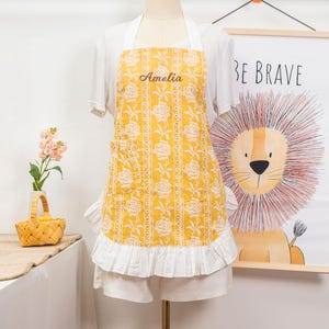 May include: Yellow apron with white rose pattern and the name "Amelia" embroidered on the chest. The apron has a white ruffle at the bottom and white straps. A poster with a lion illustration and the words "BE BRAVE" is in the background.