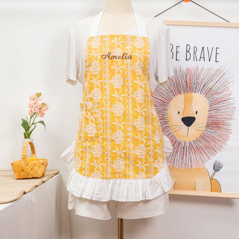 May include: Yellow apron with white rose pattern and the name "Amelia" embroidered on the chest. The apron has a white ruffle at the bottom and white straps. A poster with a lion illustration and the words "BE BRAVE" is in the background.
