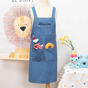Peut inclure: Tablier en jean bleu avec deux grandes poches avant et des bretelles r&eacute;glables. Le nom "Monica" est brod&eacute; en &eacute;criture noire sur la poitrine. Une souris en peluche avec un chapeau de P&egrave;re No&euml;l et un beignet jaune sont dans la poche.
