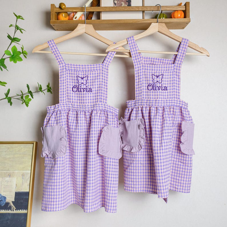 May include: Two purple and white gingham aprons with butterfly embroidery and the name "Olivia" on each. The aprons have ruffles on the pockets and are hanging on wooden hangers.
