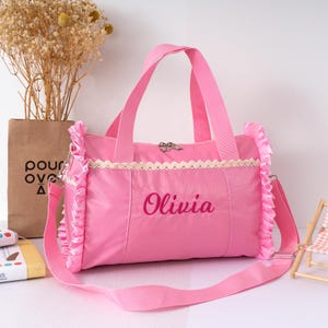 May include: A pink duffel bag with a shoulder strap and handles. The bag has a decorative lace trim and ruffled edges. The name "Olivia" is embroidered on the front. The bag is designed for carrying items.