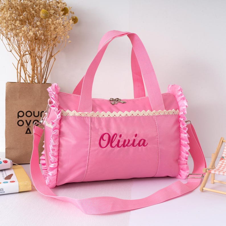 May include: A pink duffel bag with a shoulder strap and handles. The bag has a decorative lace trim and ruffled edges. The name "Olivia" is embroidered on the front. The bag is designed for carrying items.