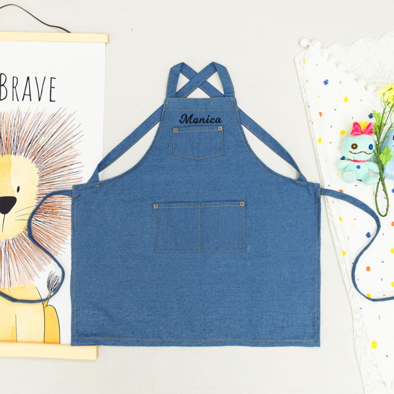 Peut inclure: Tablier en jean bleu avec le nom "Monica" brod&eacute; sur la poche de poitrine. Le tablier a deux poches inf&eacute;rieures et des bretelles r&eacute;glables. Une affiche de lion et un tissu d&eacute;coratif sont en arri&egrave;re-plan.