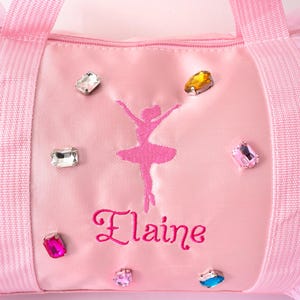 Peut inclure: Sac de sport rose avec une silhouette de ballerine brod&eacute;e en rose et le nom "Elaine". Le sac est orn&eacute; de plusieurs strass color&eacute;s. Il poss&egrave;de des poign&eacute;es roses et une fermeture &eacute;clair.