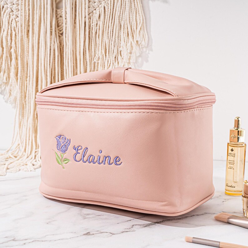 May include: A pale pink, rectangular makeup bag with a zipper closure and a bow detail. The bag has a purple embroidered rose and the name "Elaine" on the front.