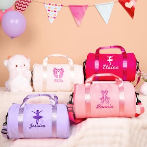 May include: Four colorful duffel bags, perfect for dance class. The bags are in white, pink, and lavender, each embroidered with a ballerina or ballet shoes and a name. The bags have handles and shoulder straps.