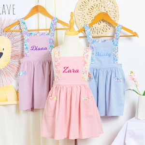 May include: Three children's pinafore dresses in pastel colors: lavender, pink, and blue. Each dress has floral patterned straps and pockets, with a name embroidered on the front. The dresses are displayed on wooden hangers.