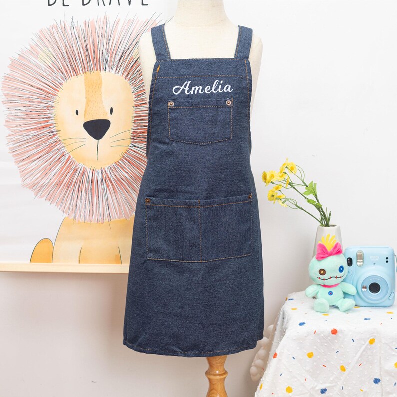 Peut inclure: Un tablier en jean bleu avec le nom "Amelia" brod&eacute; en blanc sur la poche de poitrine. Le tablier a deux grandes poches avant et des bretelles r&eacute;glables. Le tablier est pr&eacute;sent&eacute; sur un mannequin.