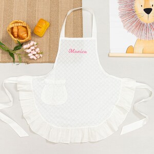 May include: White apron with a floral pattern and a ruffled hem. The apron has a pocket and the name "Monica" embroidered in pink. It has a neck strap and waist ties. The apron is displayed on a white surface.
