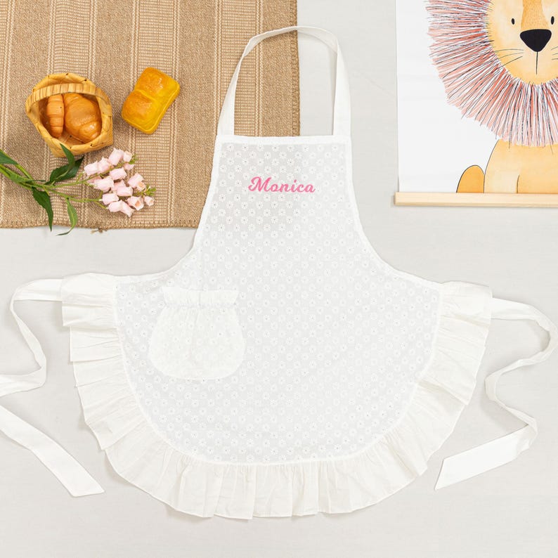 May include: White apron with a floral pattern and a ruffled hem. The apron has a pocket and the name "Monica" embroidered in pink. It has a neck strap and waist ties. The apron is displayed on a white surface.