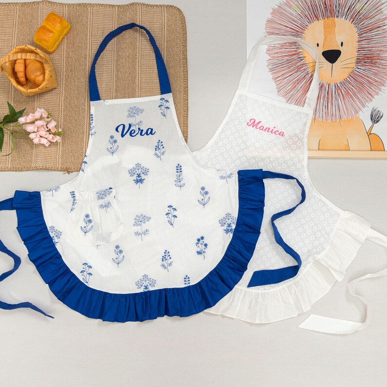 May include: Two white aprons with blue and white floral patterns and ruffles. One apron has the name "Vera" in blue script, and the other has the name "Monica" in pink script. The aprons have blue straps and ruffles.