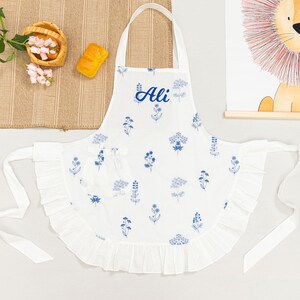 May include: White apron with blue floral pattern and the name "Ali" in script. The apron has a ruffled edge and a pocket. It is tied at the waist with long white ties. A small basket of flowers and a piece of bread are in the background.